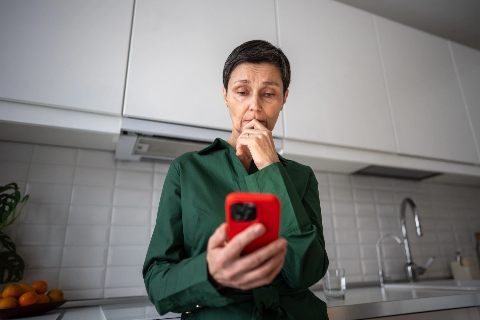 Žena v kuchyni se starostí hledí do displeje telefonu