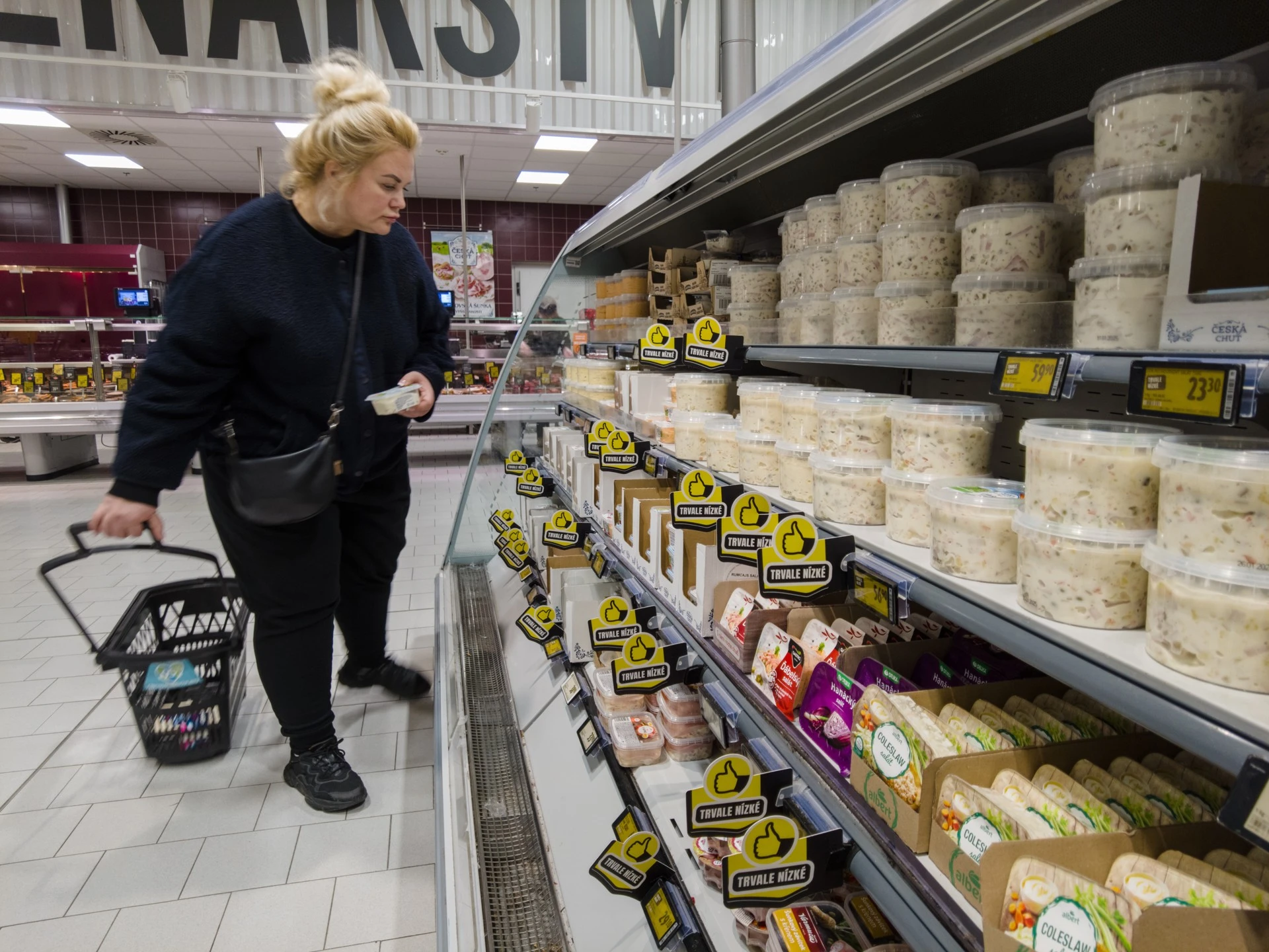 Zákaznice s košíkem u dlouhého chladicího regálu s lahůdkovými saláty v českém supermarketu.