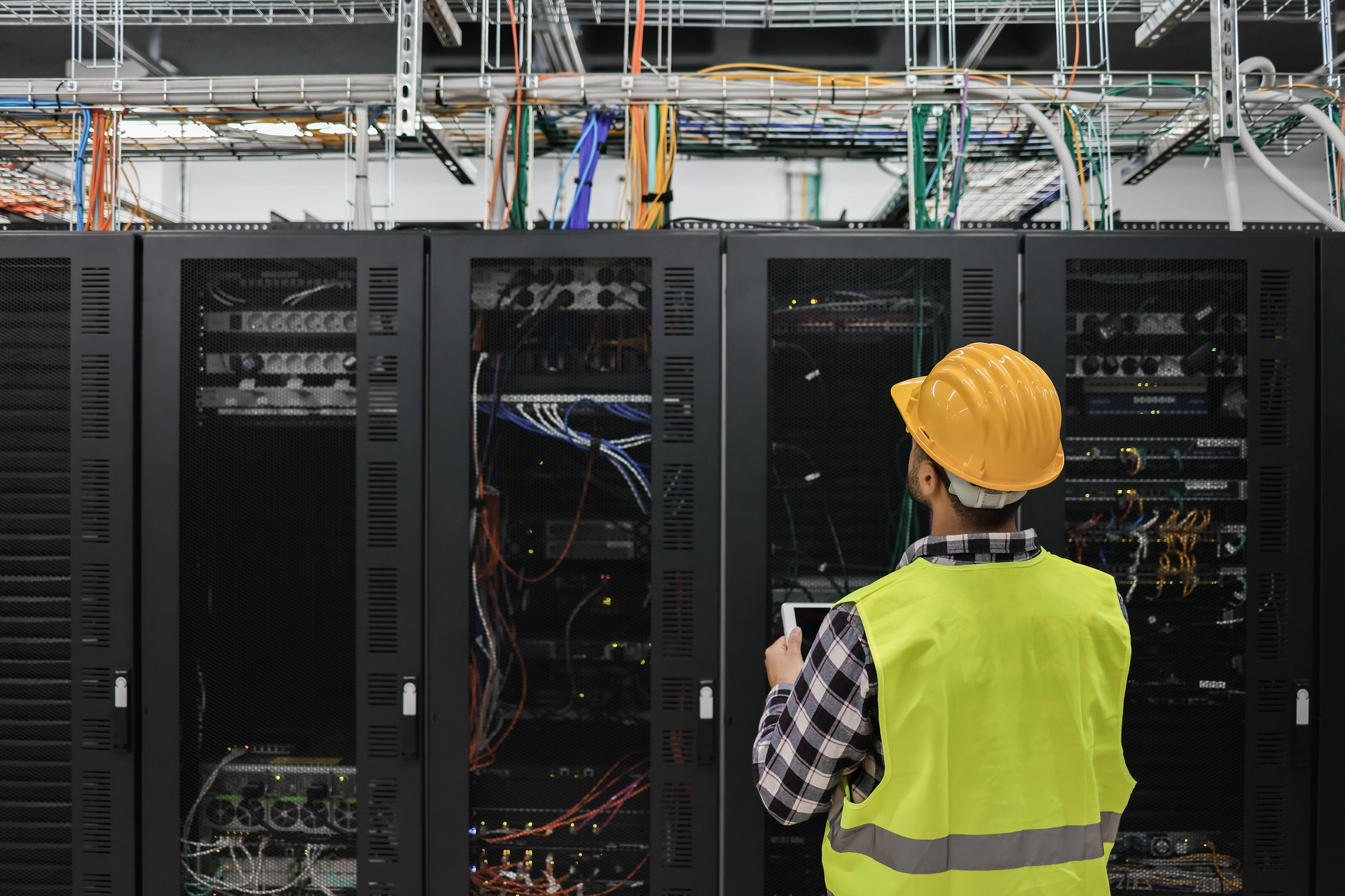 Technik v ochranné helmě a reflexní vestě stojí před řadou serverových skříní